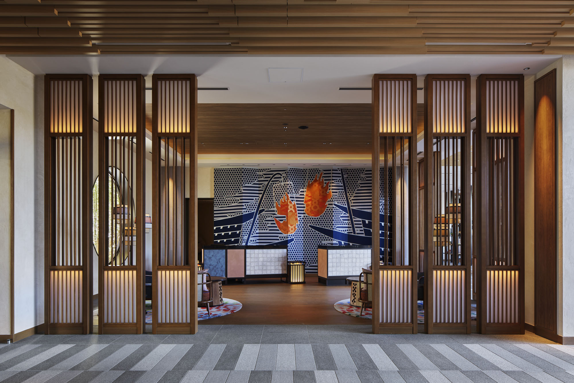 Modern hotel lobby with wooden panels and ambient lighting. A vibrant, abstract mural with bold orange shapes decorates the back wall, creating a warm, inviting atmosphere.
