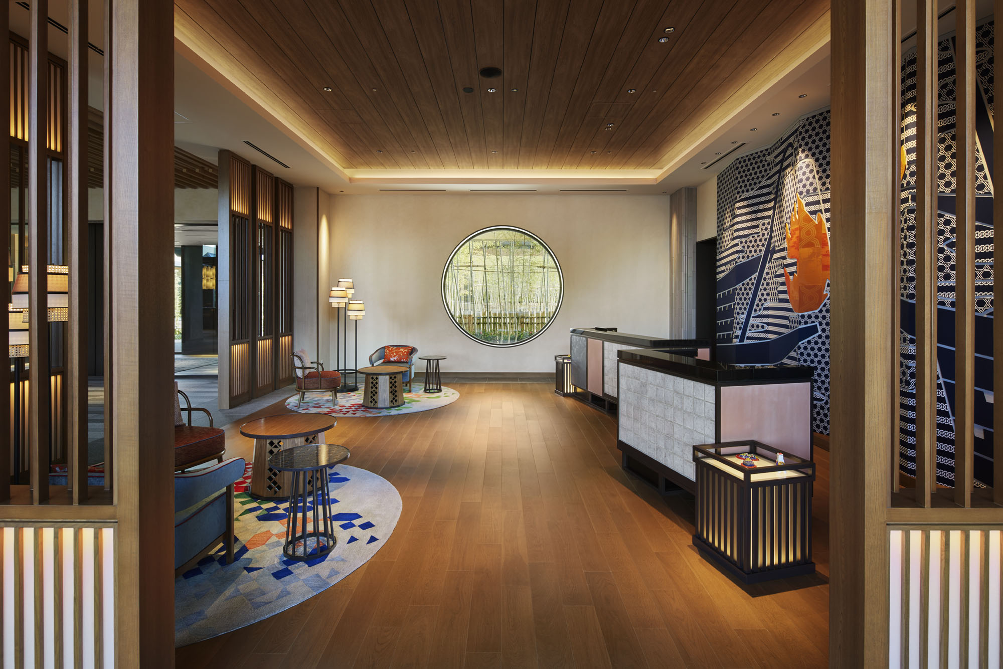 Modern hotel lobby with wooden floors, circular window with bamboo view, minimal seating, and artistic wall design creating a welcoming atmosphere.