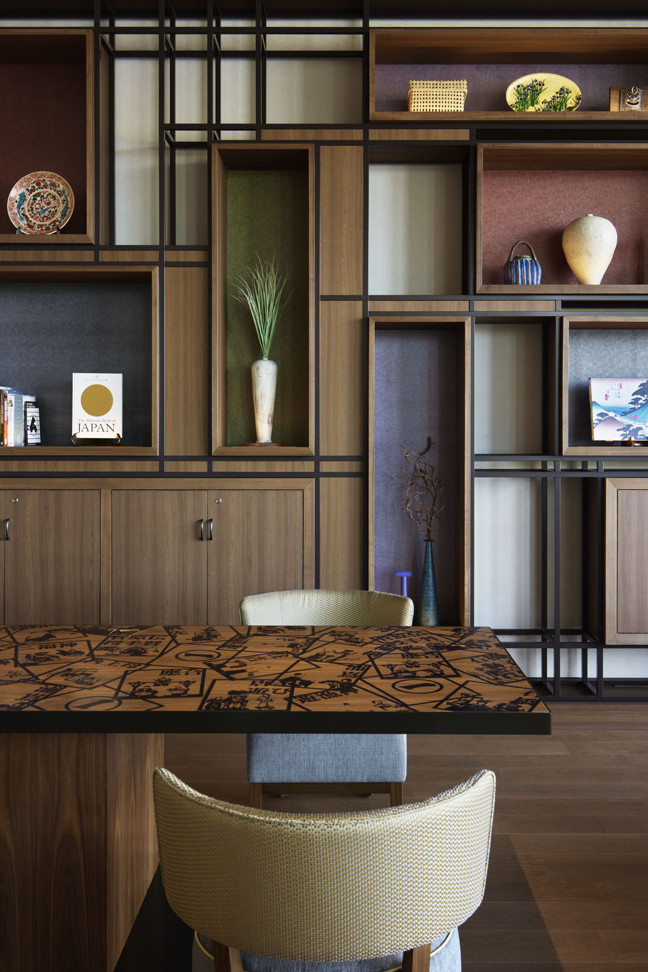 Modern hotel lobby nook with geometric wooden shelves displaying decorative items, such as ceramics and books. A patterned table and soft chair add warmth.