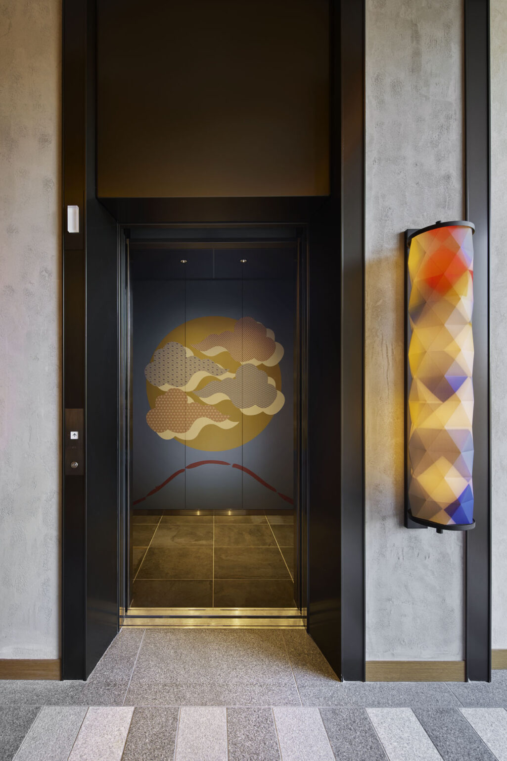 An open hotel elevator with artistic panels depicting stylized clouds and a sun against a blue background. A colorful, geometric light fixture is on the right.