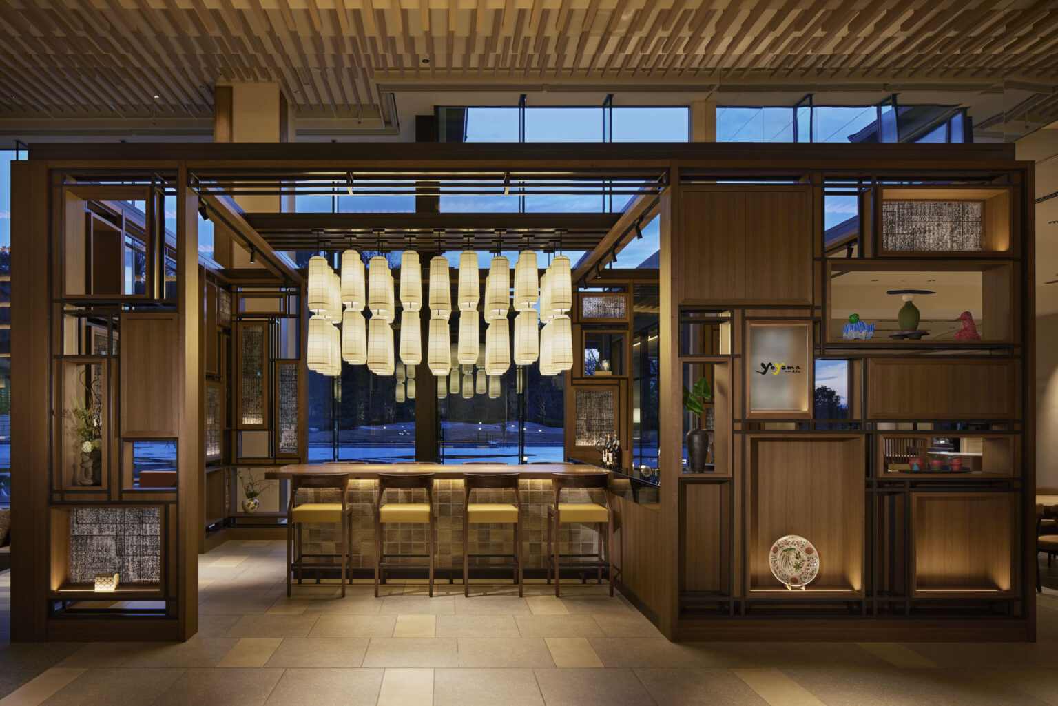 Modern hotel bar with warm lighting, featuring a row of elegant lanterns above four yellow-cushioned stools at a wooden counter. Large windows in background.