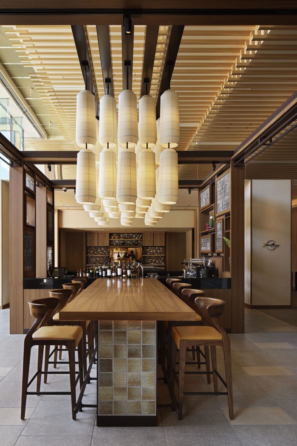 A modern hotel bar interior with a long wooden table and tall chairs. Above, soft glowing cylindrical pendant lights and wooden slatted ceiling create a warm ambiance.