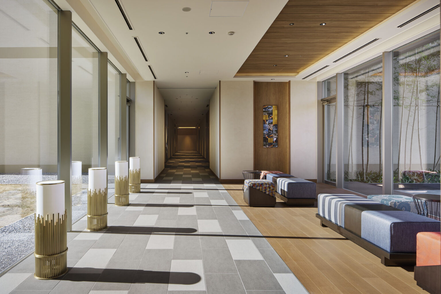 Spacious modern hotel hallway with floor-to-ceiling windows on the left, natural light illuminating patterned flooring. Soft seating on the right, serene ambiance.