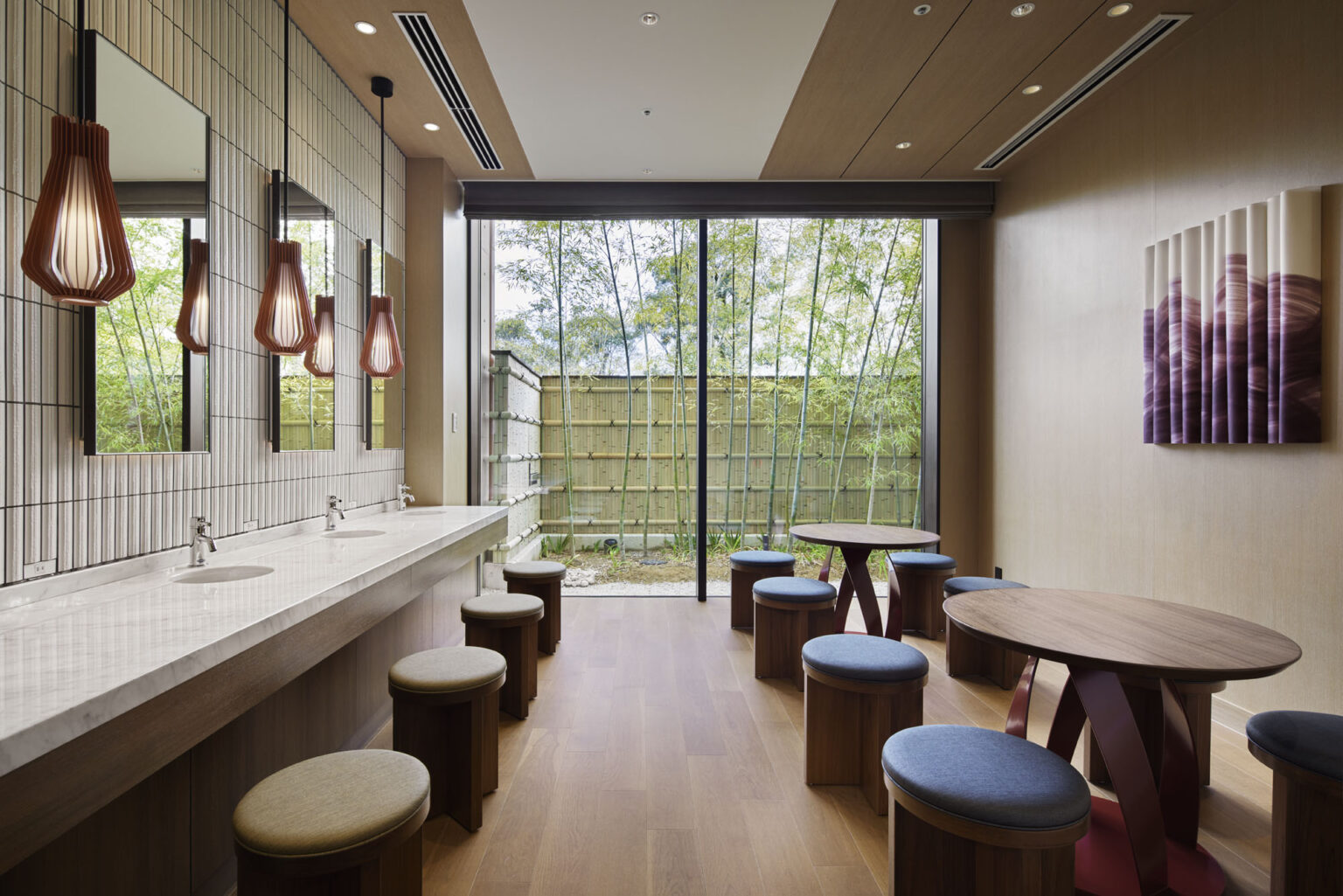 Modern hotel public restroom with wood accents, round stools, and soft lighting. Large window reveals bamboo garden, creating a serene, inviting atmosphere.