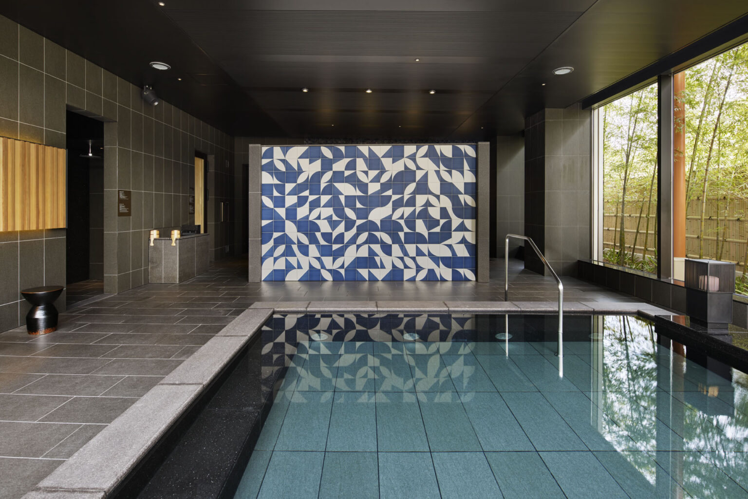 Indoor hotel pool with blue mosaic wall featuring geometric patterns, surrounded by dark tiles. Sunlight filters through large windows, creating a serene ambiance.