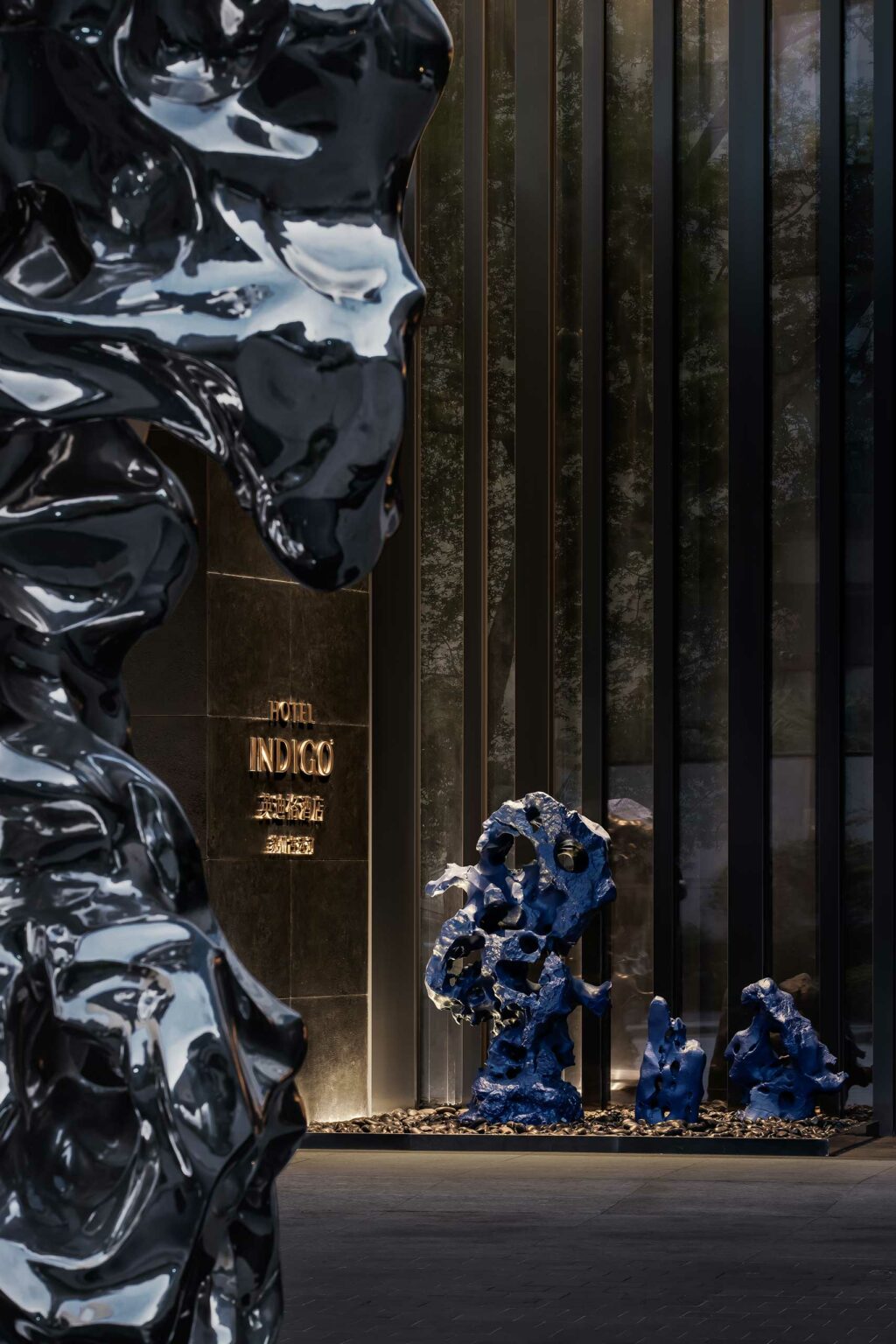 Abstract black and blue sculptures stand near a modern hotel entrance. Soft lighting casts reflections, creating an elegant, contemporary atmosphere.