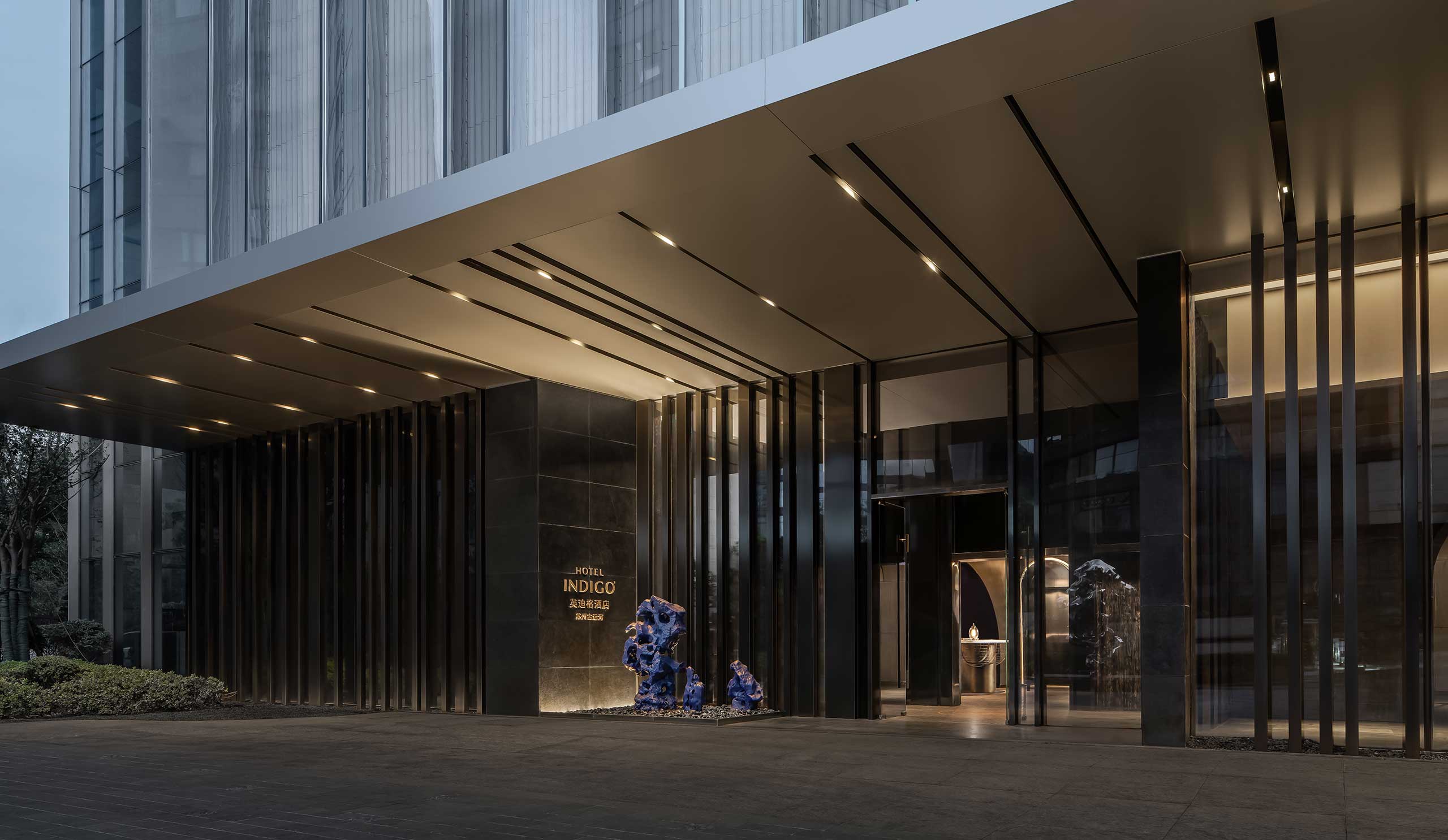 Modern hotel entrance featuring a sleek, minimalist design with dark panels and vertical lines. Blue sculptures add a bold contrast, creating a sophisticated ambiance.