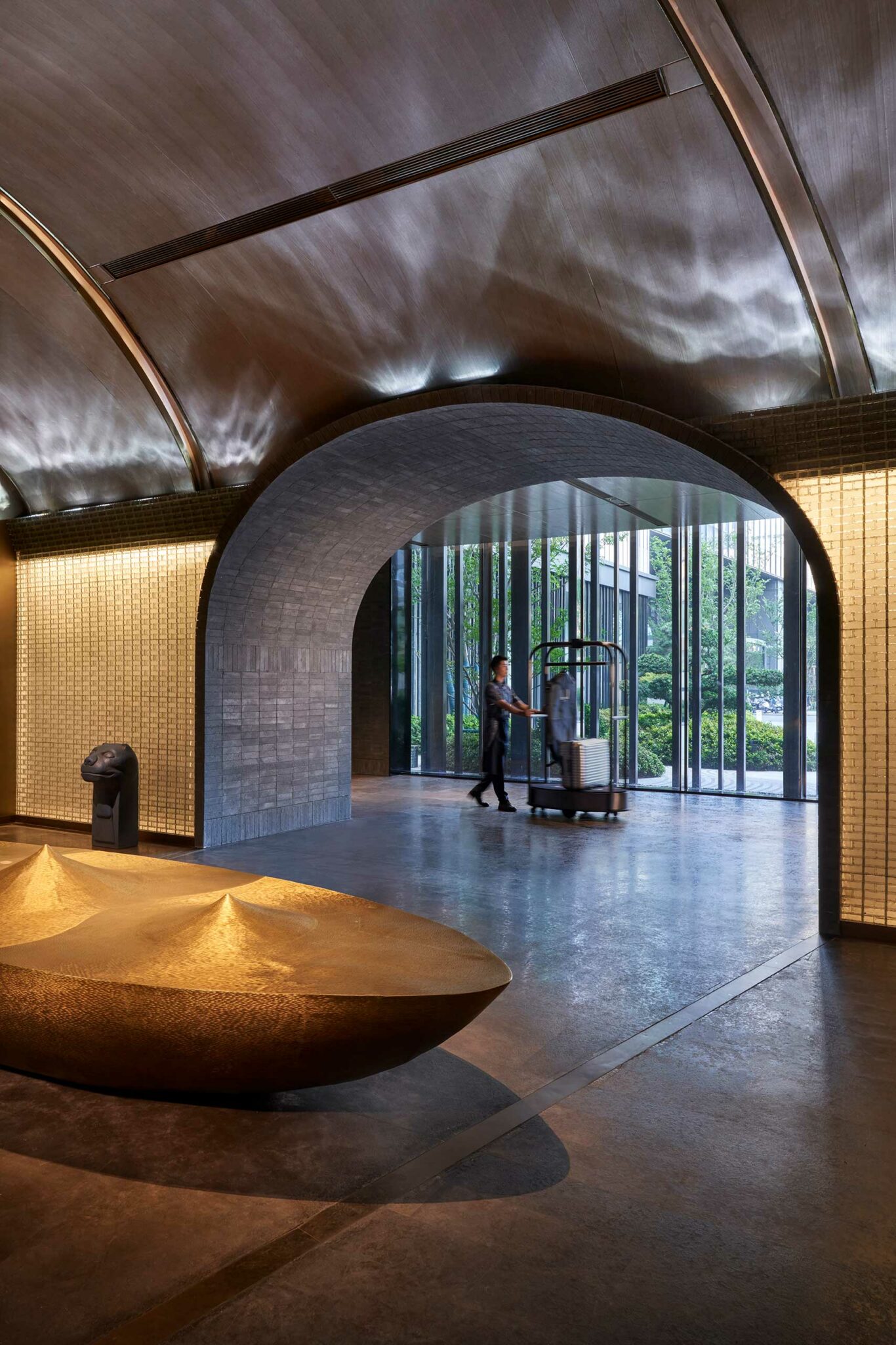 A modern hotel lobby with a vaulted ceiling, a person pushes a luggage cart through a stone archway, large windows reveal greenery outside. Calm atmosphere.