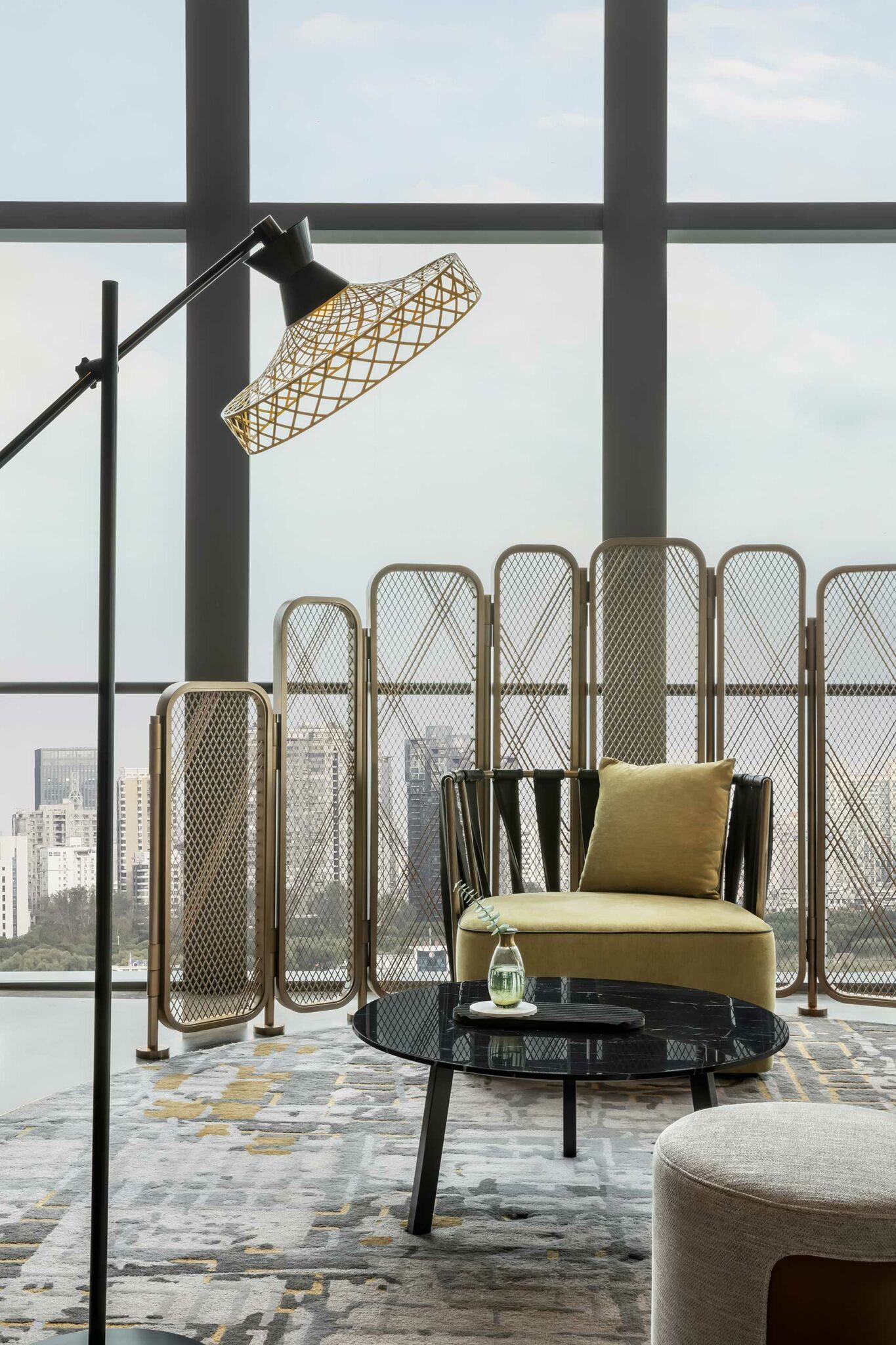 Modern hotel lounge nook with a large window revealing a cityscape. Features a floor lamp, metallic divider, yellow chair, pillow, black coffee table, and glass vase.