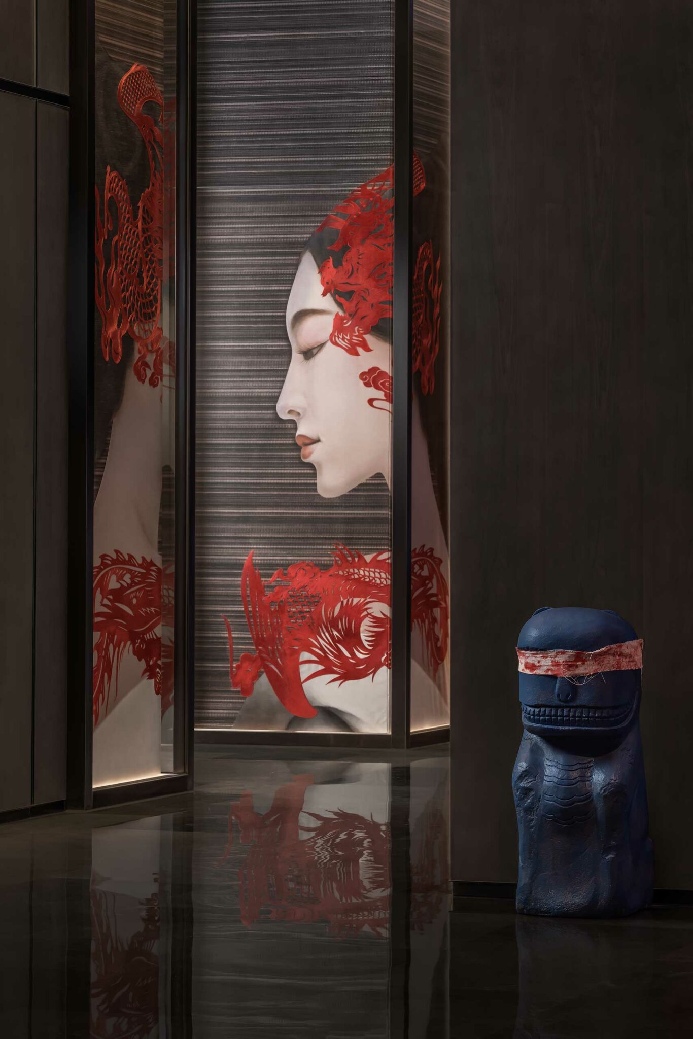 A serene profile of a woman with red floral patterns on a panel is reflected in a polished floor. A small blue sculpture stands nearby, adding contrast.