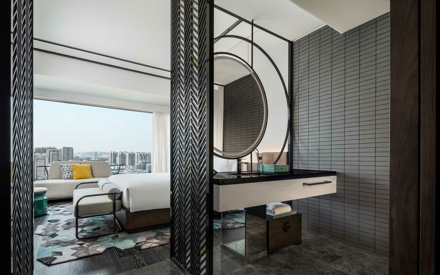 Modern hotel room with a city view. Features a double bed, a circular mirror above a black countertop, and a gray brick accent wall, conveying sleek elegance.