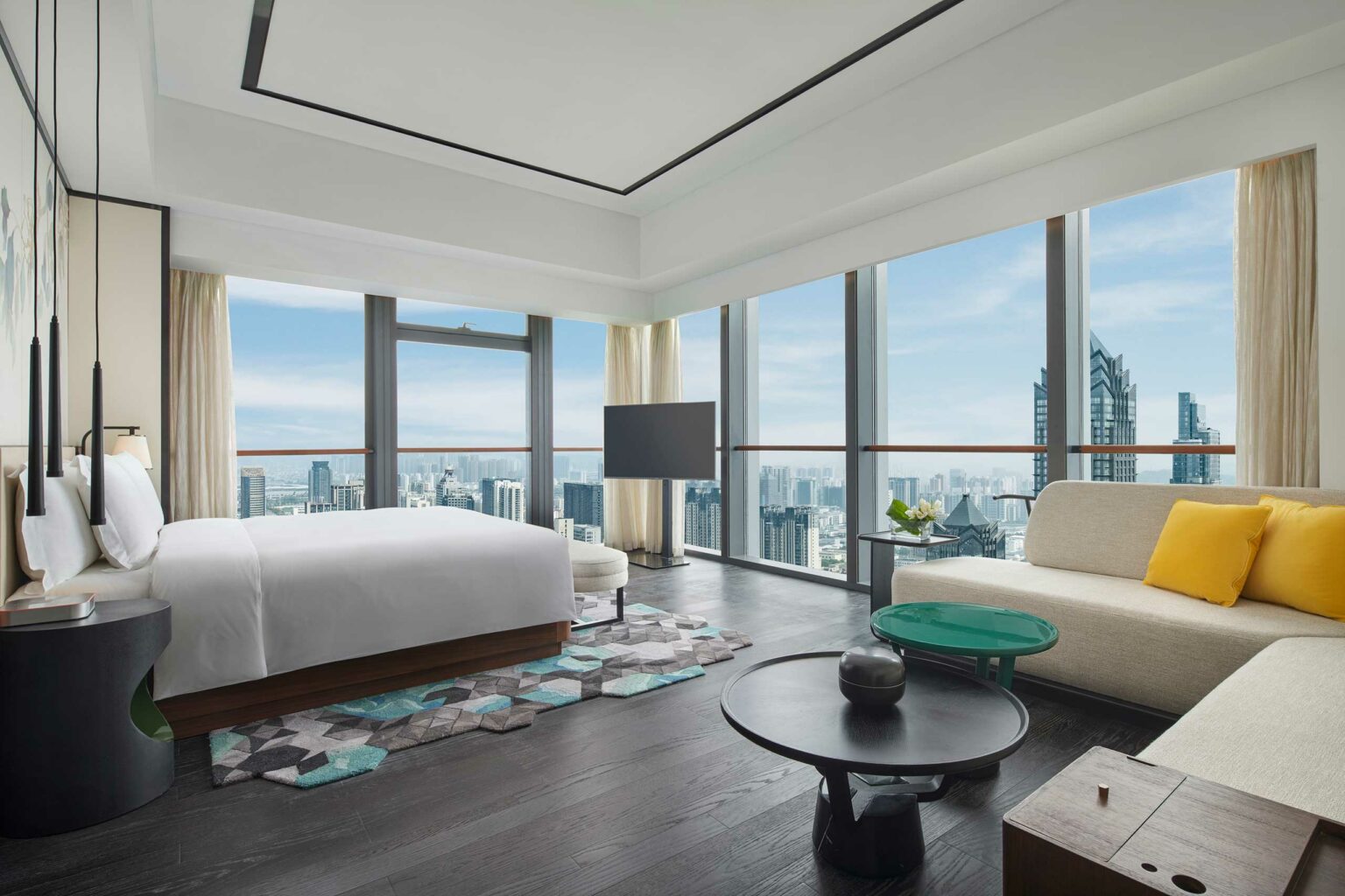 Luxurious hotel room with floor-to-ceiling windows showcasing a stunning cityscape. Features a plush bed, modern sofa with yellow pillow, and sleek decor.