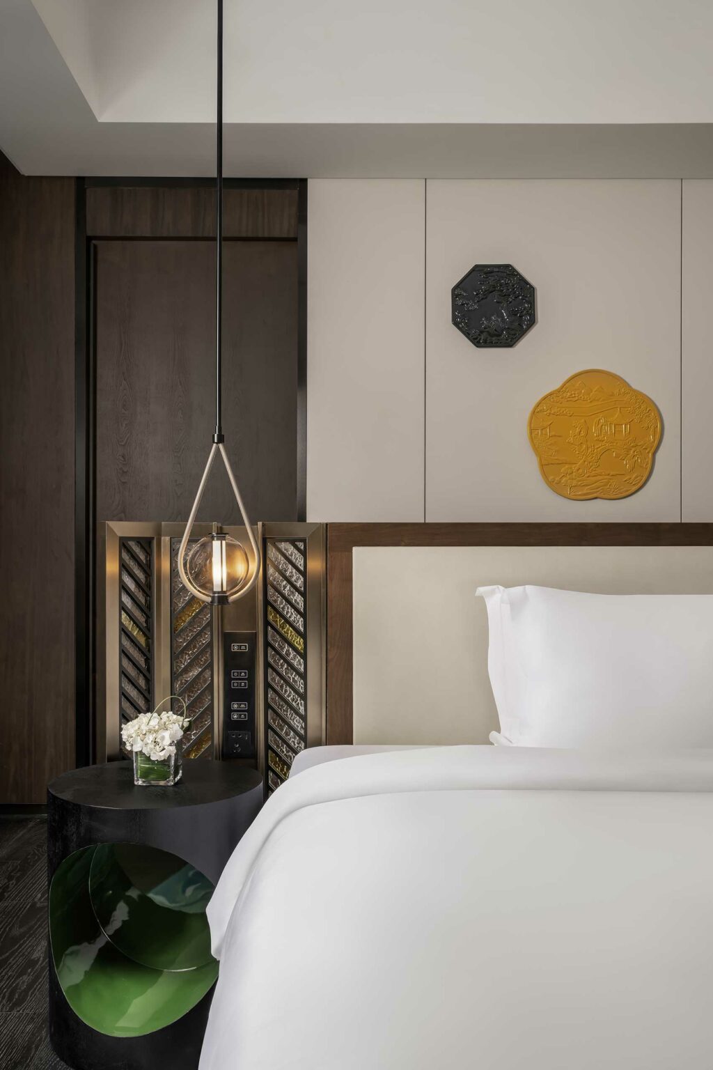 Modern hotel room with a luxurious white bed, a minimalist pendant light, and a round black table holding white flowers. Neutral tones add a serene feel.