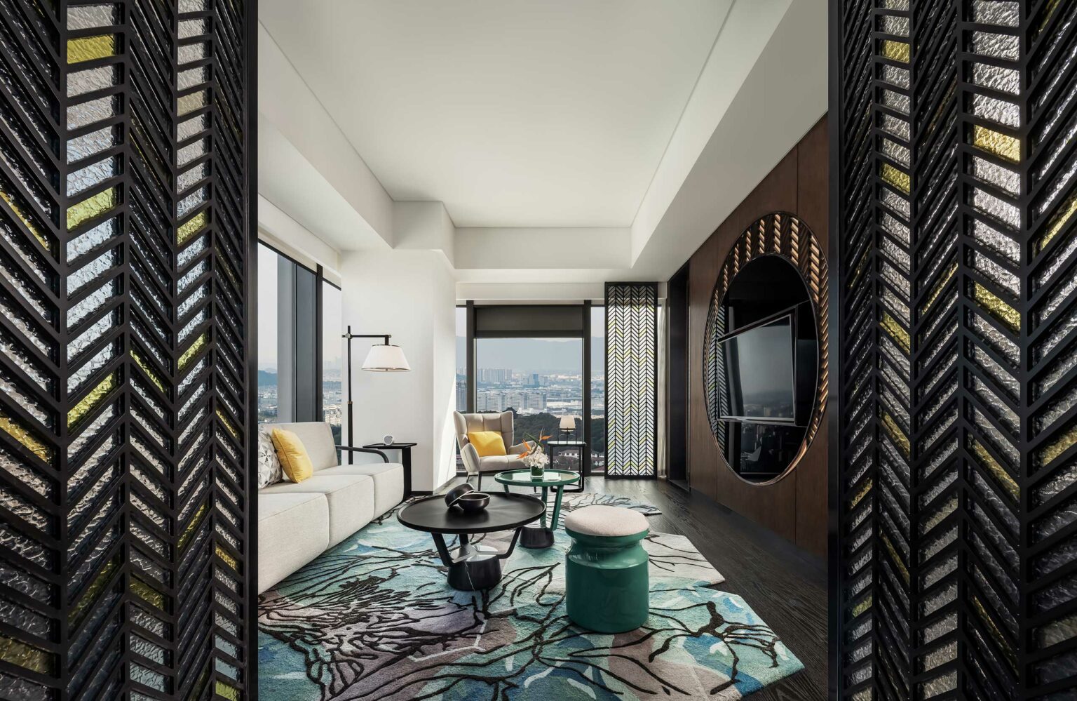 Modern guestroom living room with a large window view of the city. It features a white sofa, yellow cushions, round tables, a patterned rug, and a wall TV.