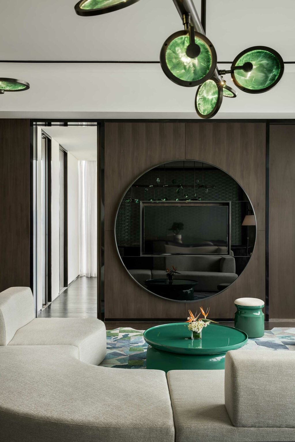 Modern guestroom living room with a circular mirror on a dark wood wall, reflecting a couch and lamp. Features a green glass chandelier and matching round table, neutral-toned sofa, and geometric rug. Calm and contemporary.