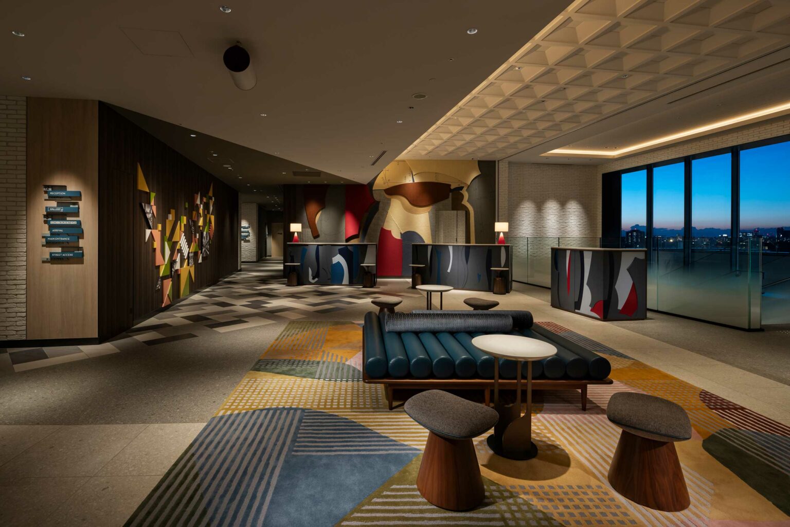 Modern hotel lobby featuring abstract wall art, geometric-patterned carpet, and stylish seating. Large windows reveal a cityscape at dusk, creating a sophisticated ambiance.
