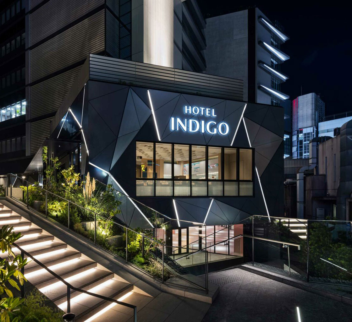 Night view of Hotel Indigo Tokyo Shibuya's modern exterior, featuring angular panels and illuminated signage. Steps with soft lighting lead up, surrounded by greenery.