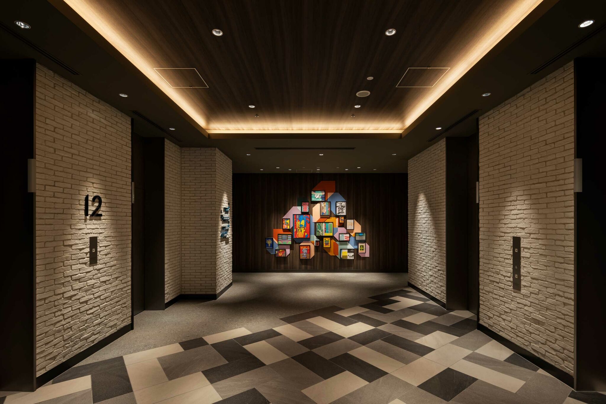 Dimly lit hotel lift lobby with a modern art display featuring colorful frames on a dark wooden wall. Brick walls and patterned floor tiles add texture.