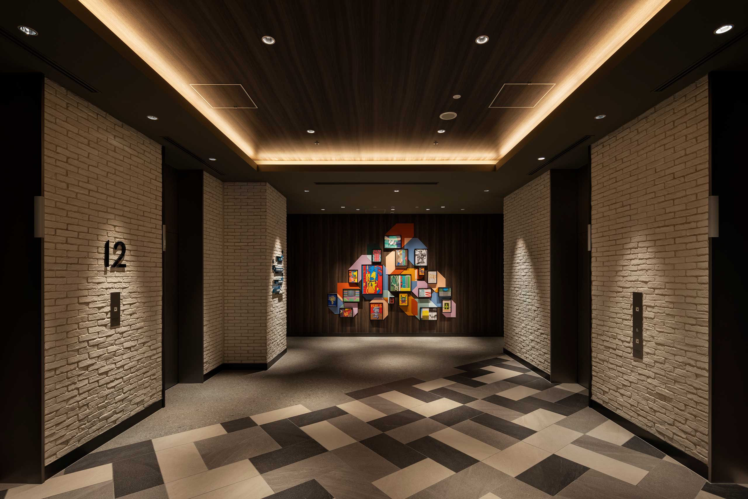Dimly lit hotel lift lobby with a modern art display featuring colorful frames on a dark wooden wall. Brick walls and patterned floor tiles add texture.