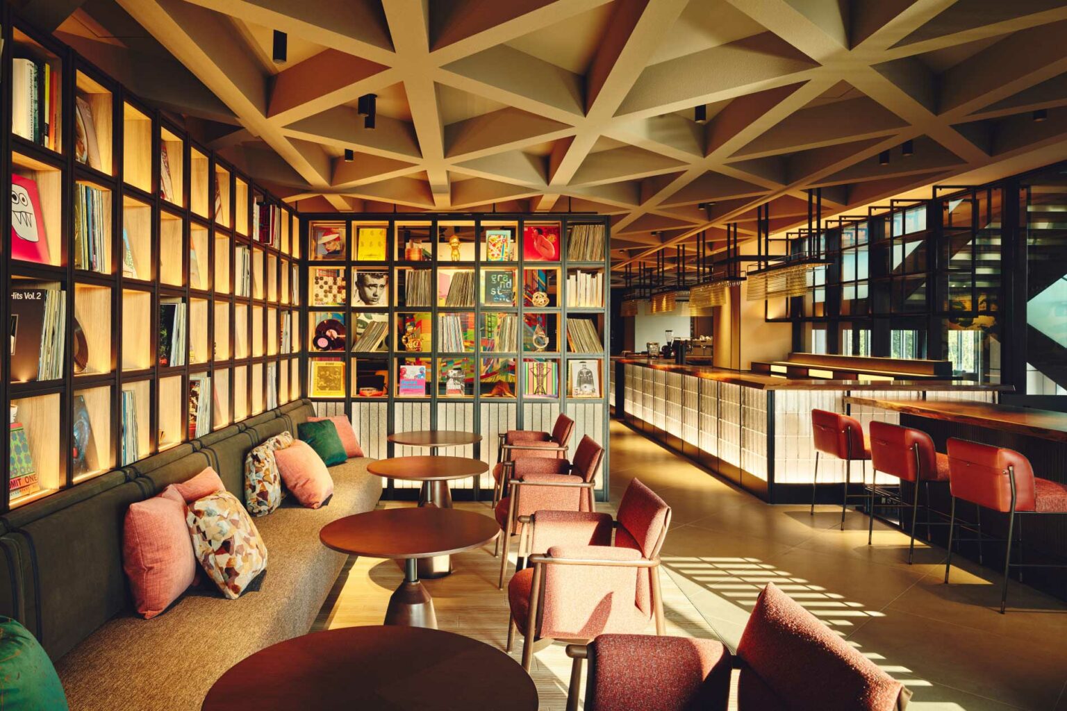 Modern hotel lounge with geometric ceiling, vinyl records on shelves, cozy seating with colorful pillows, and a sleek bar, creating a warm, inviting ambiance.