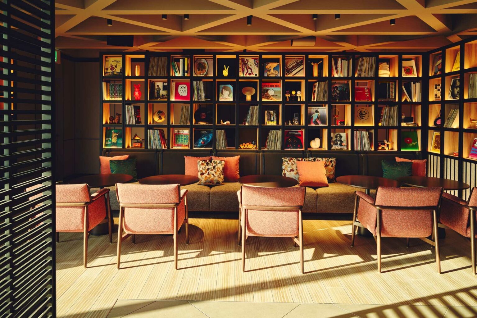 Cozy hotel lounge with warm lighting, featuring a wall of neatly arranged books and records. Four armchairs face a sofa adorned with colorful cushions.
