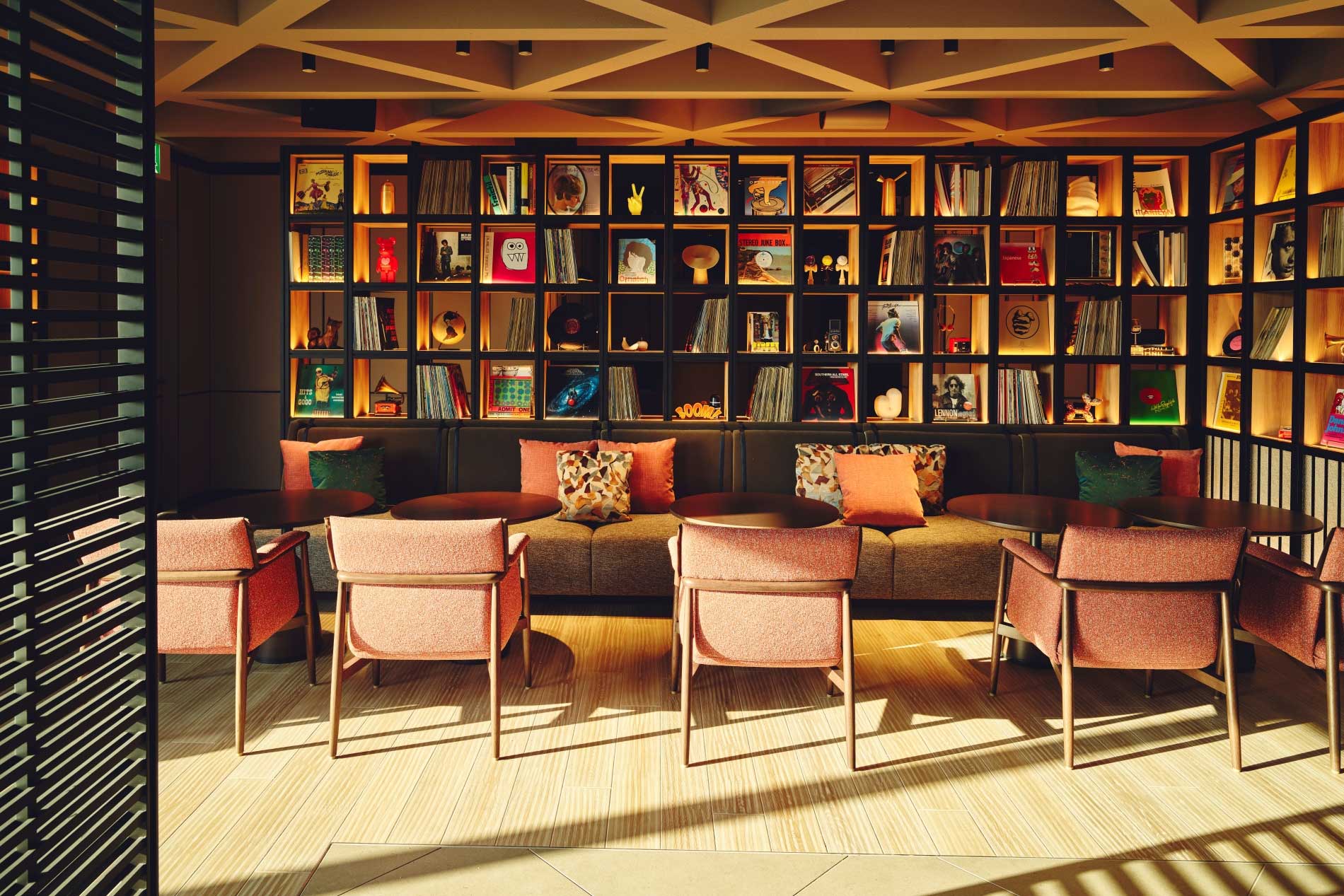 Cozy hotel lounge with warm lighting, featuring a wall of neatly arranged books and records. Four armchairs face a sofa adorned with colorful cushions.