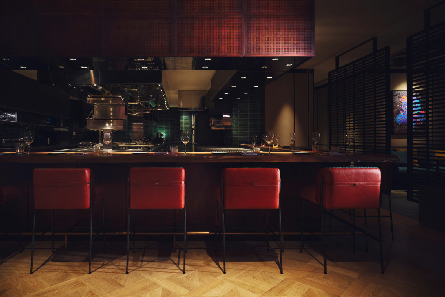 Dimly lit hotel restaurant bar with a warm, cozy ambiance. Five red leather bar stools line the counter, with glassware sparkling under soft lighting.