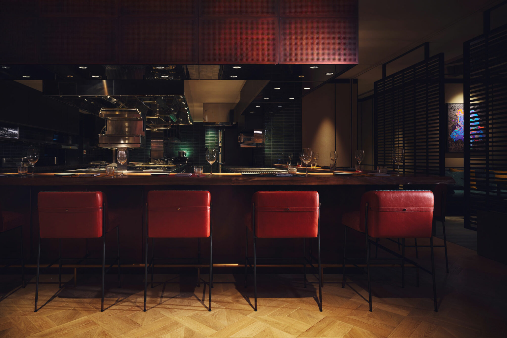 Dimly lit hotel restaurant bar with a warm, cozy ambiance. Five red leather bar stools line the counter, with glassware sparkling under soft lighting.