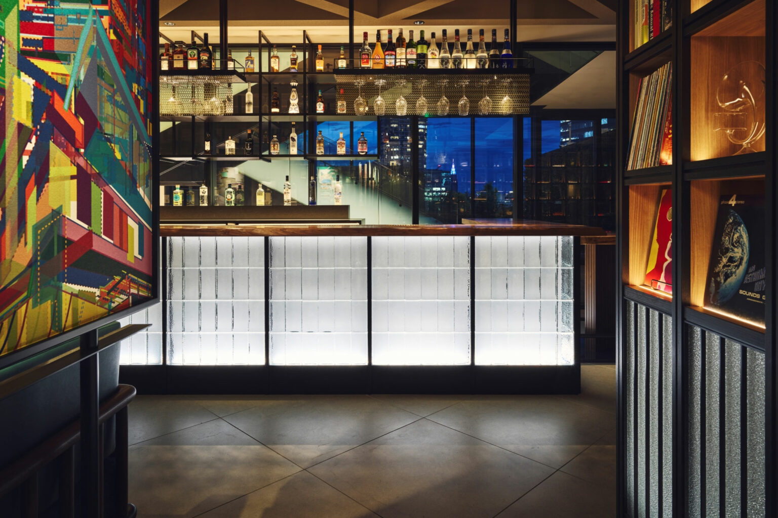 Stylish hotel bar interior with illuminated counter, colorful abstract art on the wall, and bottles on glass shelves. City skyline visible through window.