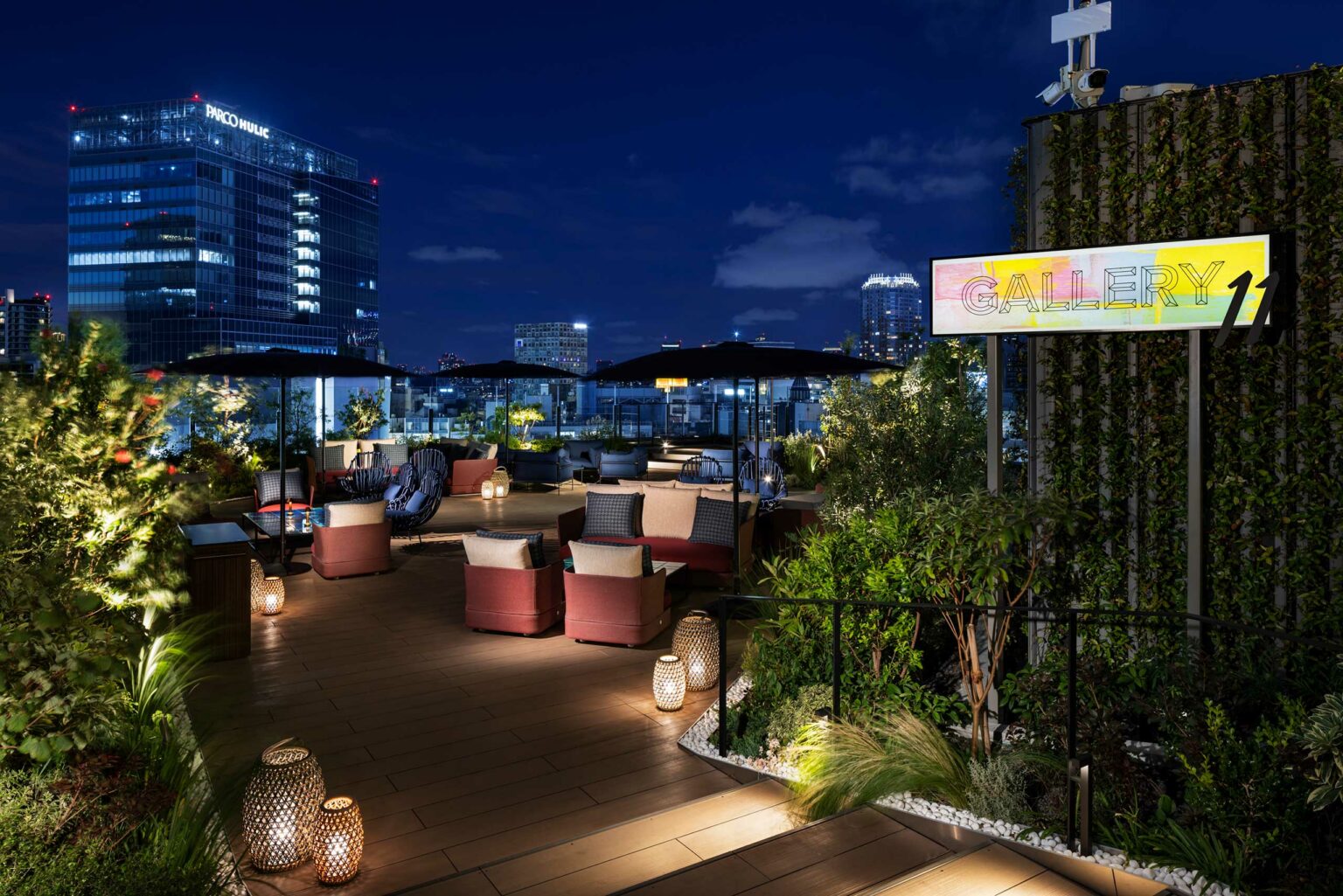 Rooftop lounge at night with cozy seating, soft lighting, and plants; a cityscape backdrop under a starry sky. Neon
