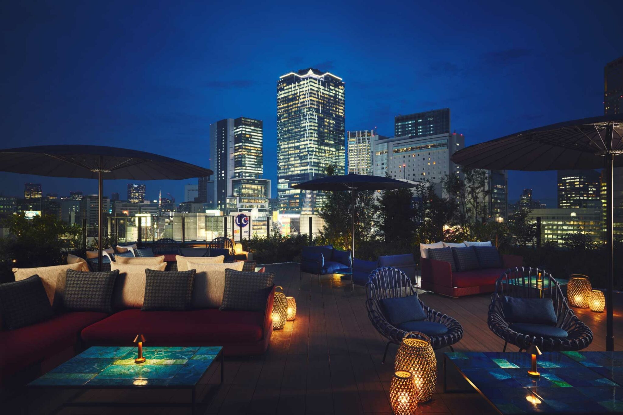 A rooftop lounge at night, featuring modern seating with lanterns casting a warm glow. A city skyline with illuminated skyscrapers forms the backdrop.