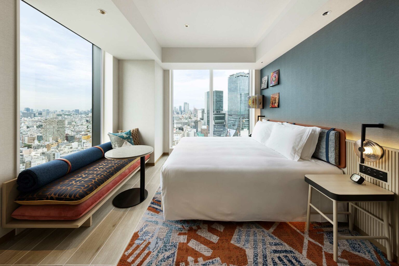 Modern hotel room featuring a large bed, stylish bench with cushions, and floor-to-ceiling windows offering a panoramic city skyline view. Serene and inviting ambiance.