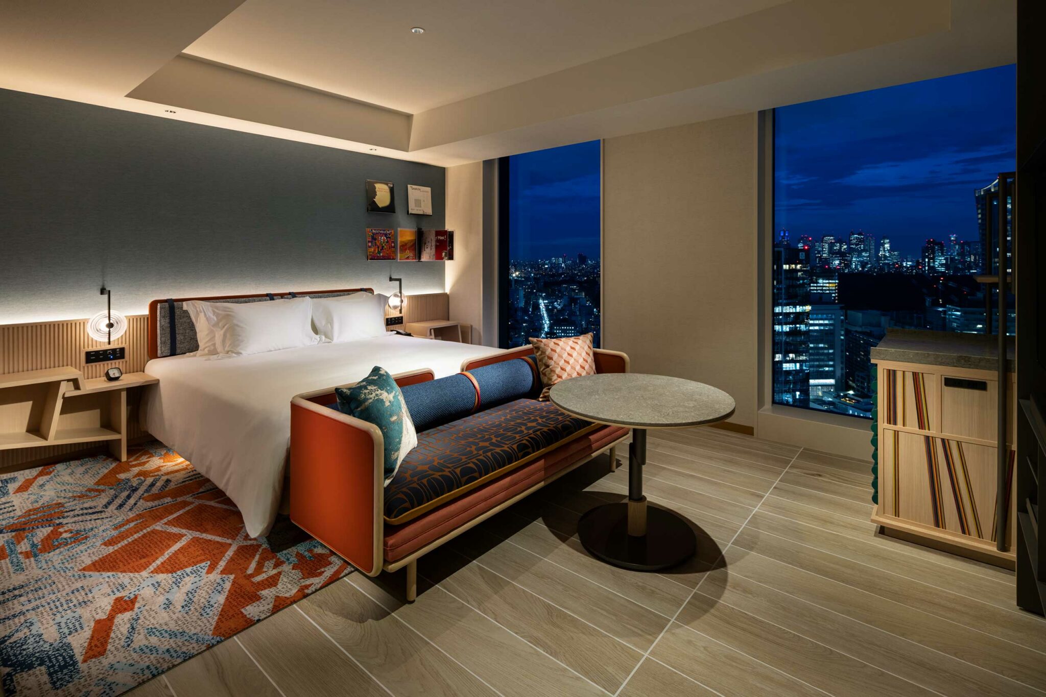 Modern hotel room with a large bed, elegant sofa, and round table. Night cityscape visible through a floor-to-ceiling window, creating a cozy, luxurious ambiance.
