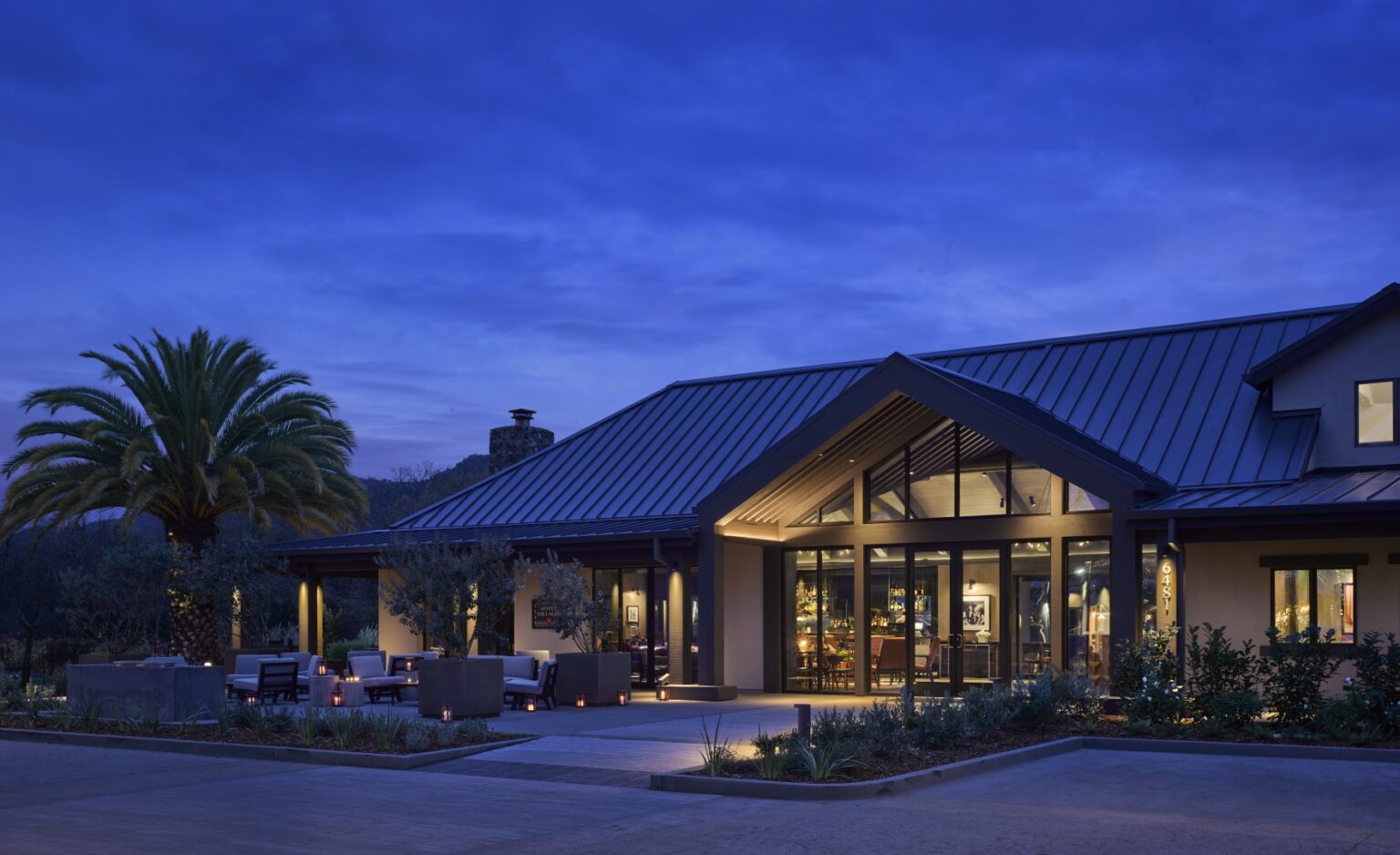Exterior of Hotel Villagio Yountville Napa featuring a modern building at dusk with warm interior lights glowing through large windows. A palm tree and patio seating create a tranquil, inviting atmosphere.