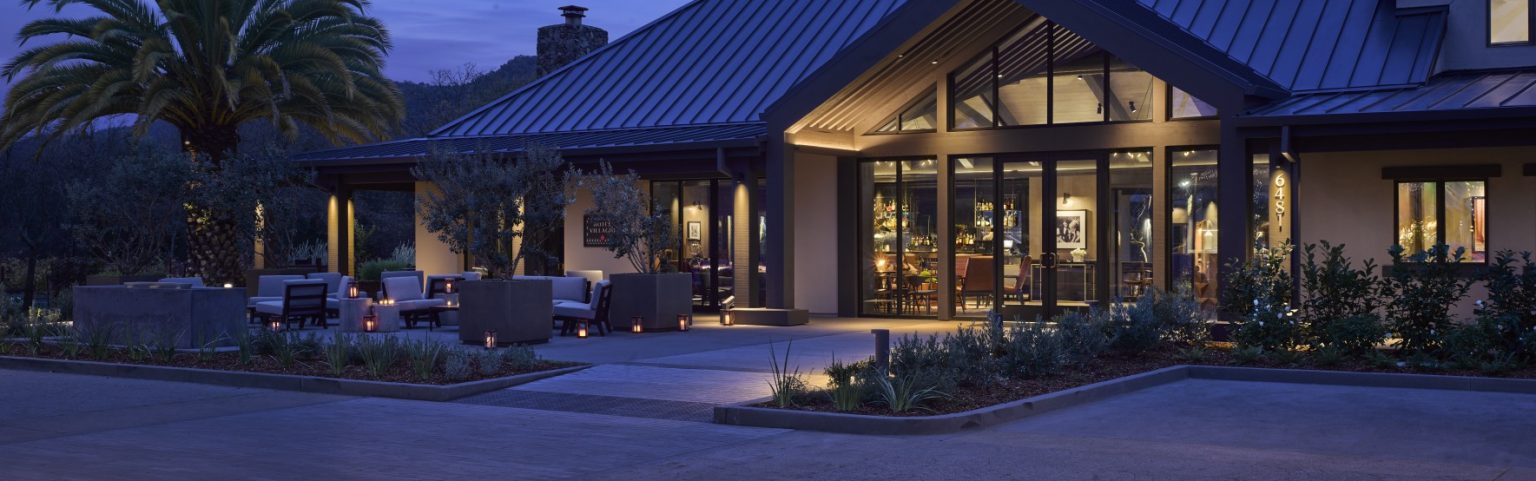 Exterior of Hotel Villagio Yountville Napa featuring a modern building at dusk with warm interior lights glowing through large windows. A palm tree and patio seating create a tranquil, inviting atmosphere.
