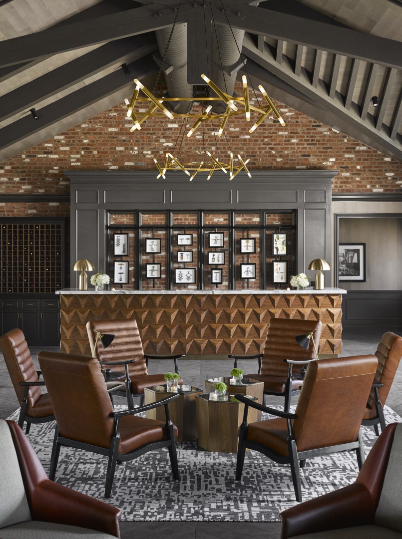 Contemporary hotel lounge with leather chairs facing a geometric wooden bar. Brick wall with framed art behind. Modern chandelier adds elegance and warmth.