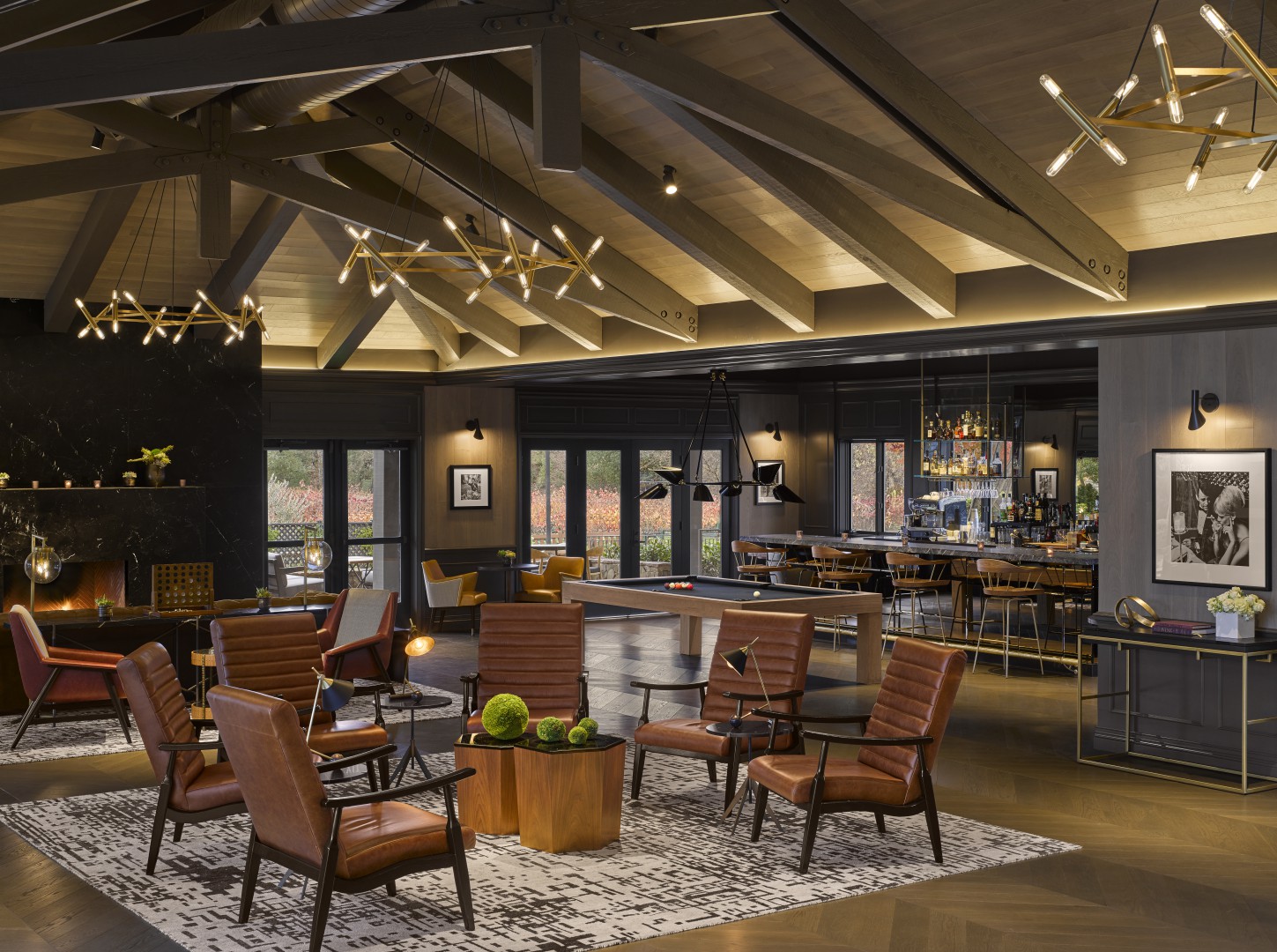 Modern hotel lounge with leather chairs, wood tables, and geometric chandeliers. Features a bar, pool table, cozy fireplace, and ceiling beams, exuding warmth.