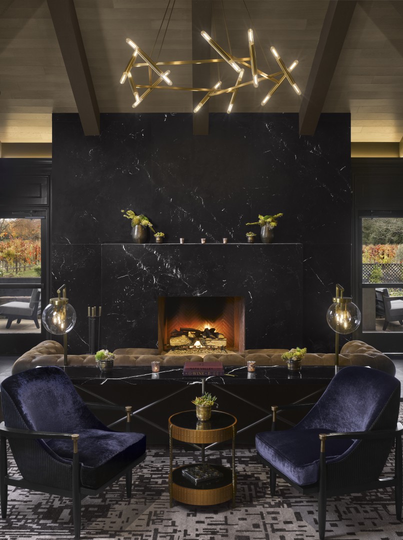 Luxurious hotel lounge with a dark marble fireplace, warm fire, modern chandelier, two blue velvet chairs, and gold-accented decor creating a cozy ambiance.