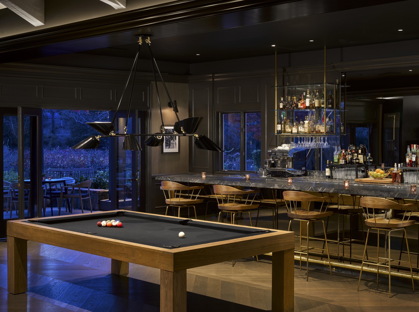 Stylish hotel bar interior with a modern pool table in the foreground, dim lighting, wooden chairs, stocked bar, and a serene landscape visible through large windows.