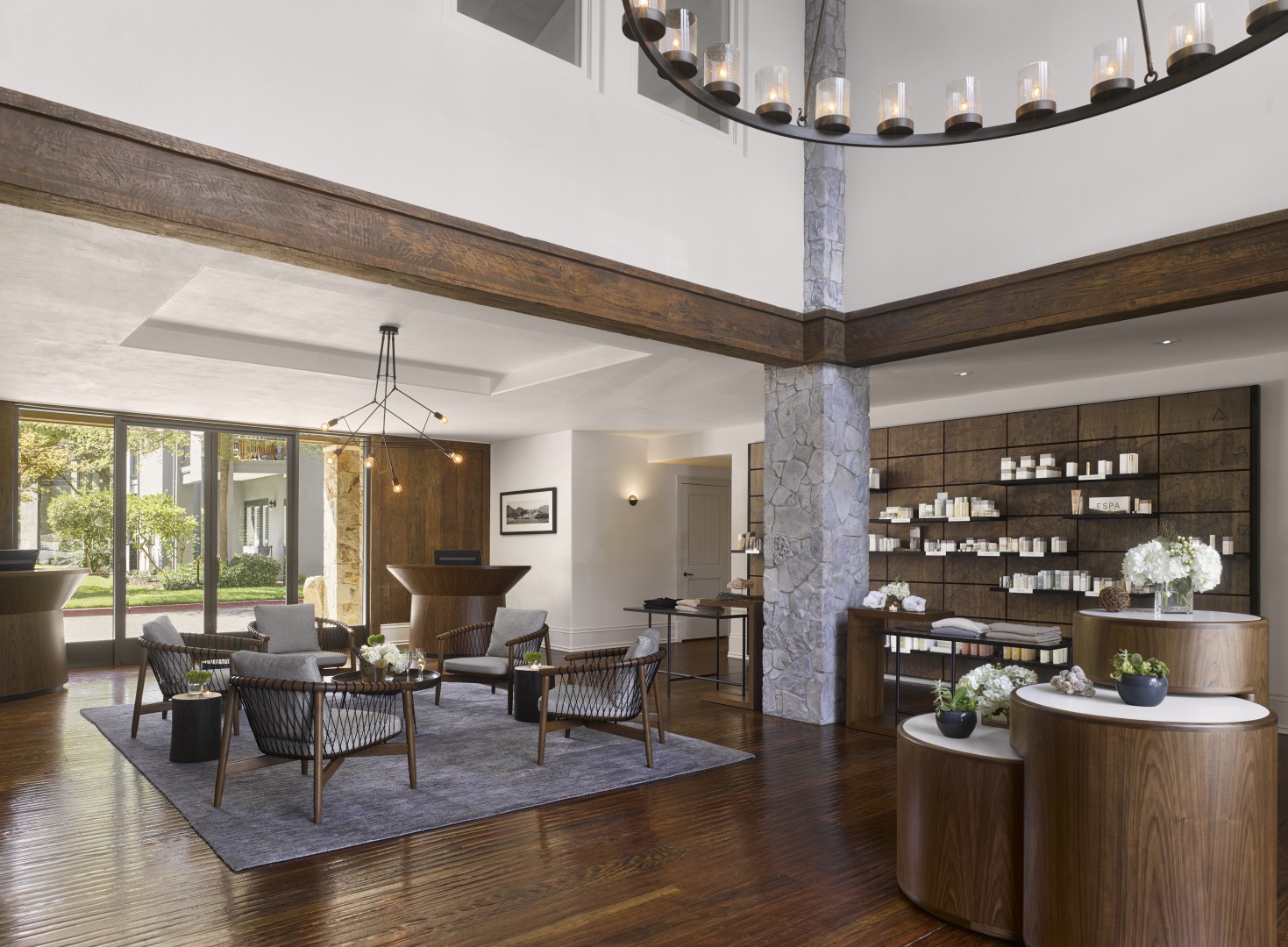 Spacious, modern hotel lobby with wood accents, a large circular chandelier, and elegant seating. Bright natural light and shelves with cosmetics create a serene atmosphere.