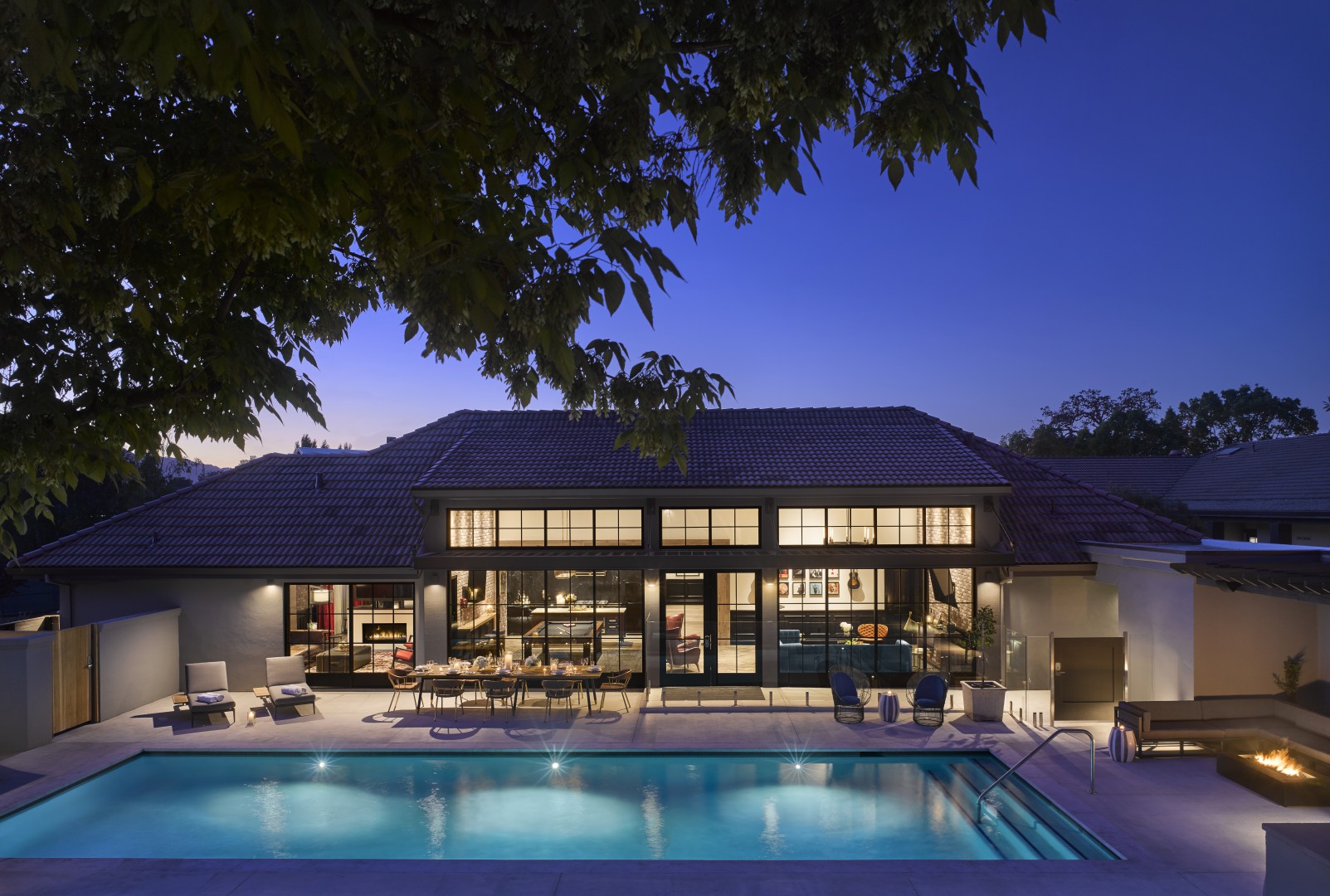 Modern hotel building at dusk, with large windows softly illuminated. A swimming pool and patio furniture create a tranquil and inviting atmosphere.