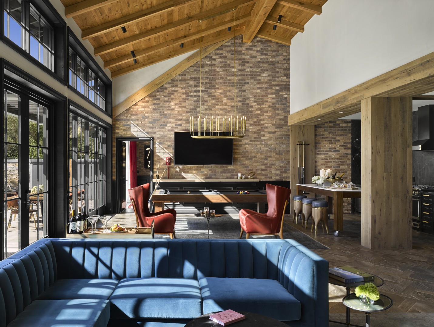 Spacious sunlit hotel lounge with a vaulted wood ceiling, brick wall, and large windows. Features a pool table, modern chandelier, red chairs, and a blue sofa.