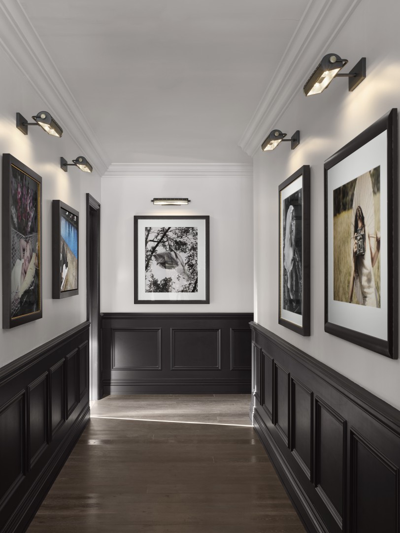 A sleek hotel hallway features black-and-white framed art on white walls, complemented by dark paneling. Overhead lights create a sophisticated ambiance.