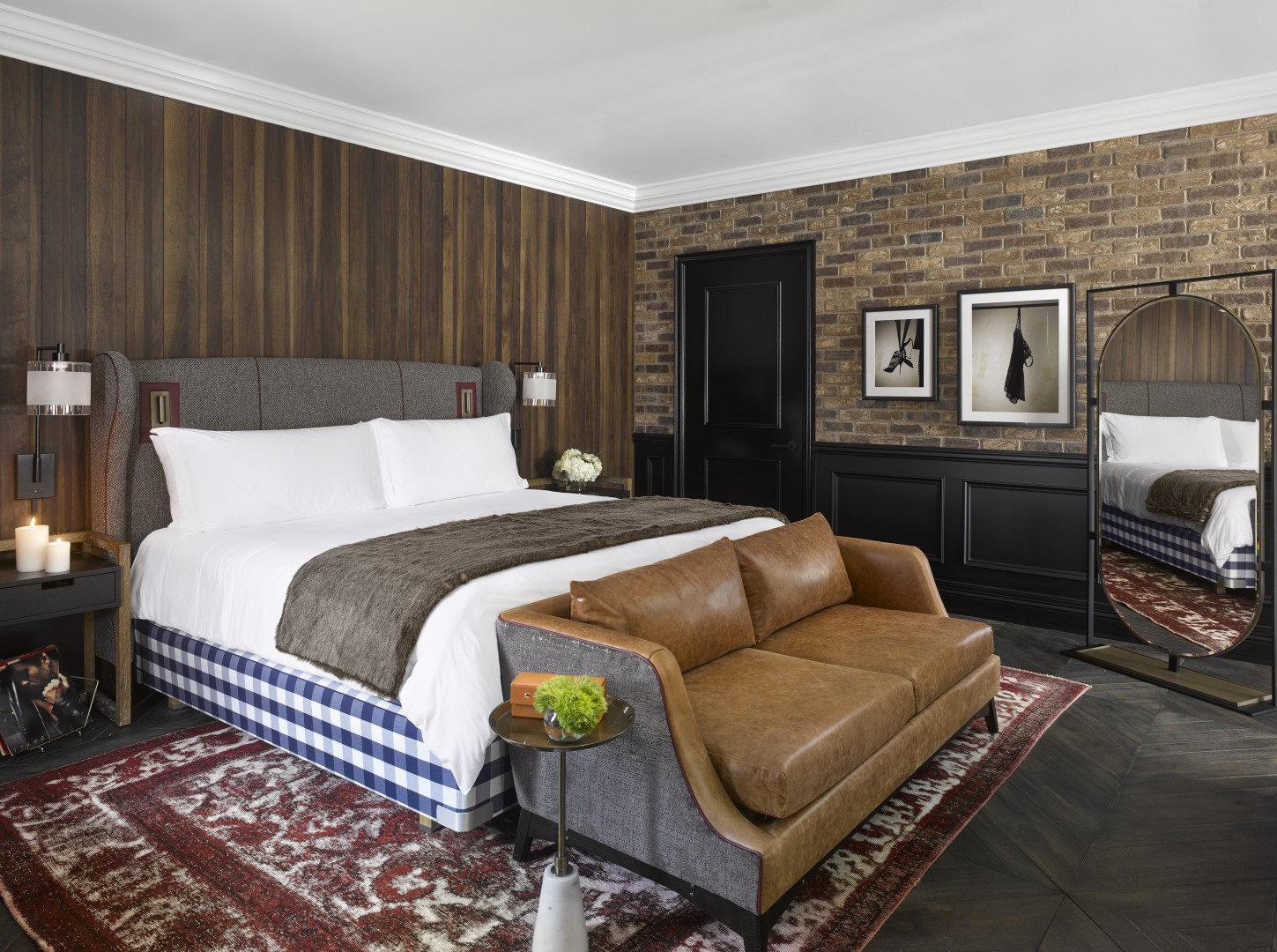 Stylish hotel room with a large bed, white linens, and gray headboard. A brown leather sofa sits at the foot, with modern decor and warm lighting.