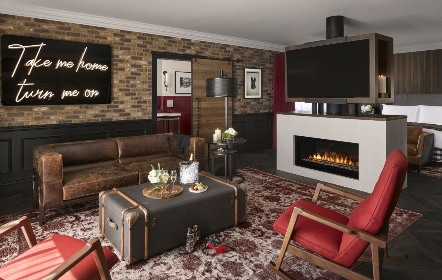 Stylish suite living room with a brown leather couch, vibrant red chairs, a trunk-style coffee table, fireplace, and neon sign reading, 