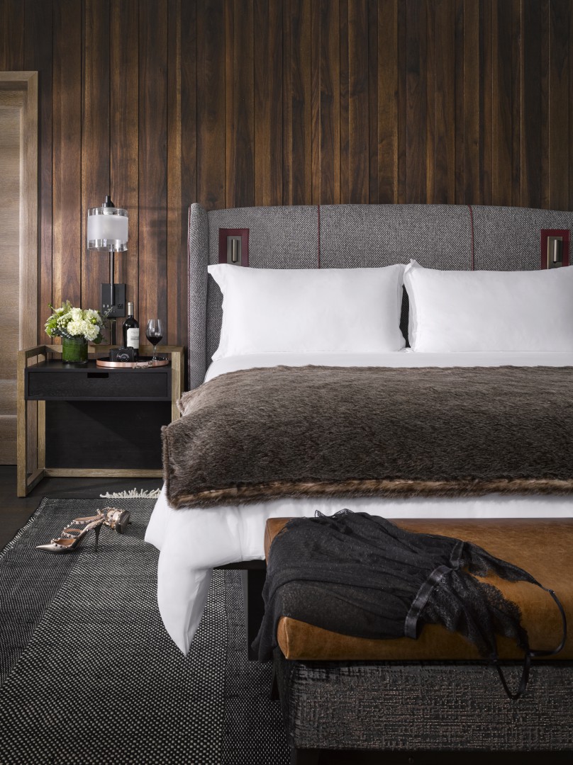 Luxurious hotel bedroom with a wood-paneled wall, plush bed with white pillows, fur throw, and bedside table with flowers, wine, glasses, and a lamp. Cozy and elegant.