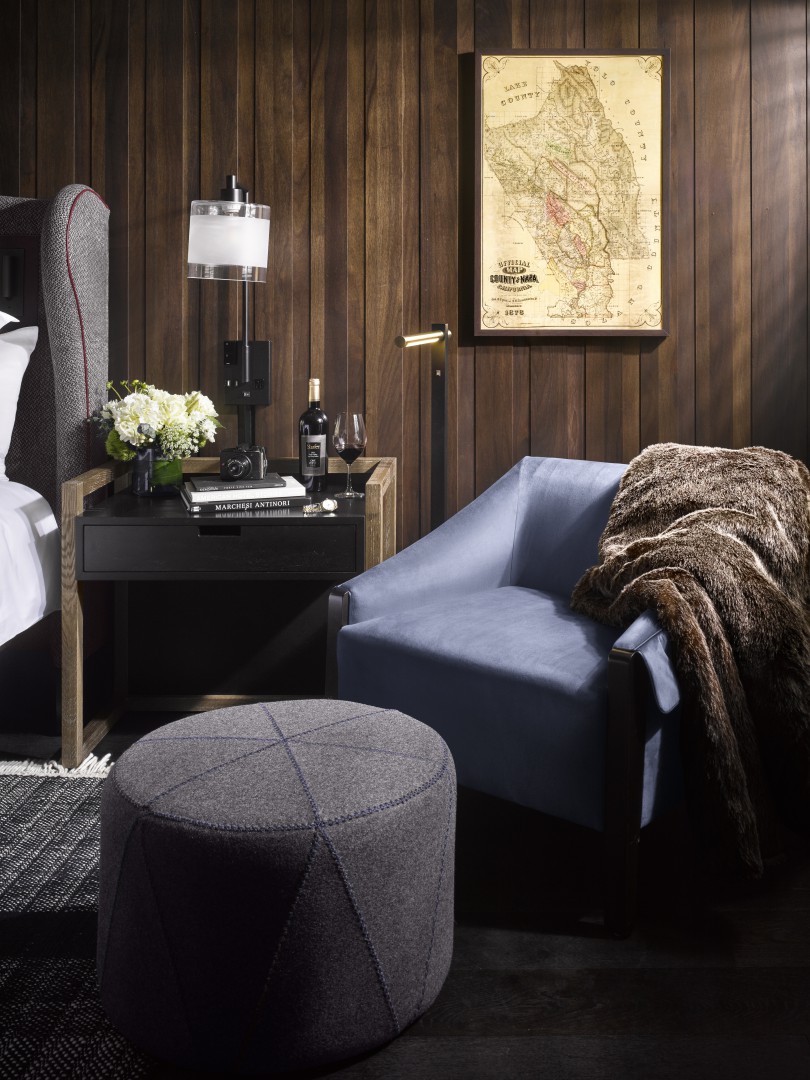 Cozy hotel room with a soft blue armchair draped with a fur throw, wooden walls, a vintage map, a stylish side table, and a grey pouf. Warm and inviting.
