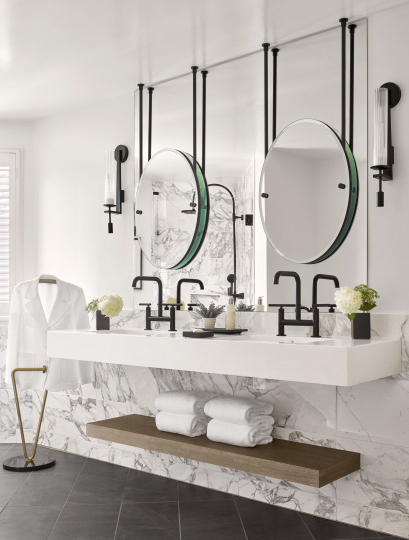 Elegant suite bathroom with marble walls and counter, twin black faucets, round mirrors, and modern sconces. White towels and robe create a serene tone.