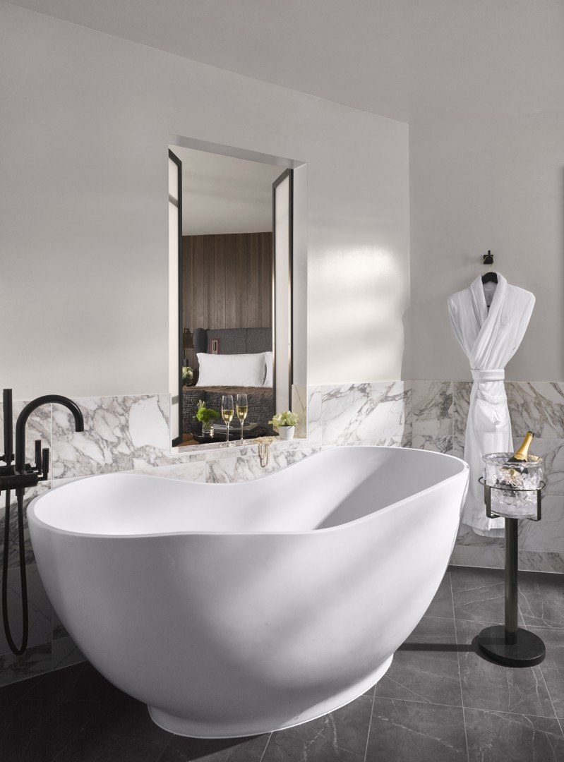 Spacious, modern suite bathroom with a large white freestanding tub, marble walls, black fixtures, a hanging robe, and a window view into a cozy bedroom.