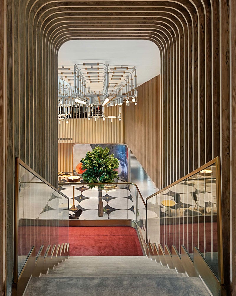 Elegant staircase with wooden slats leads down to a modern lobby with geometric floor patterns, lush green plant, and unique overhead light fixture.